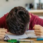 A person is sitting at a table with their head resting on an open notebook, looking frustrated. Various pens and a pencil are scattered on the table.